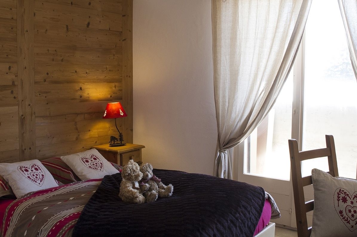 A cozy bedroom with a wooden headboard, a bed with heart-patterned pillows, a bedside lamp, and a window with sheer curtains.