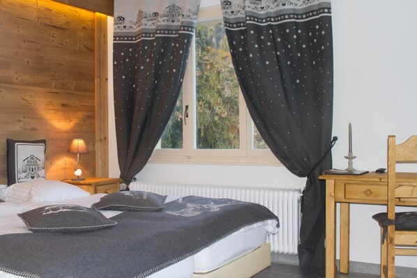 A bedroom with a wooden headboard, a bed with a dark blanket, two pillows, a bedside table with a lamp, and a window with patterned curtains. - Hotel-Restaurant de la Poste (Ayent)