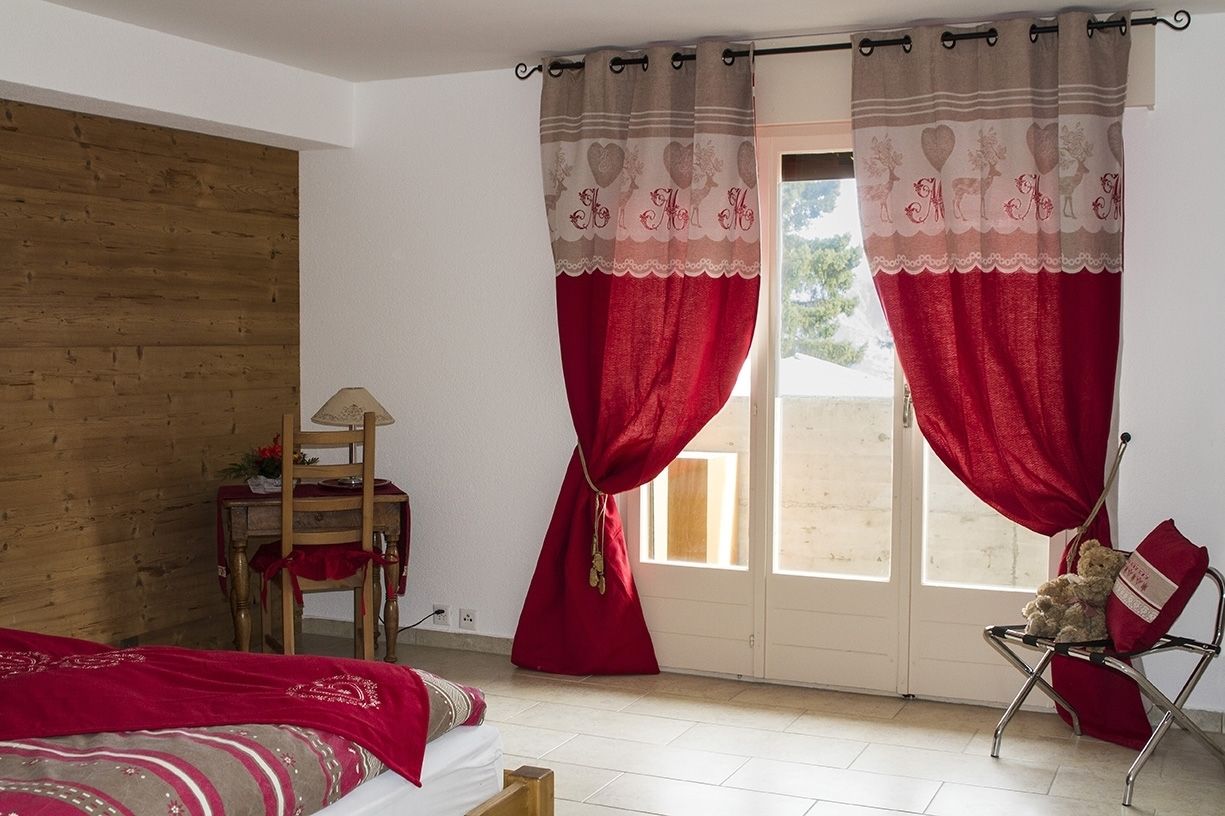 Un dormitorio con pared de madera, una cama, un sillón con un oso de peluche y una ventana con cortinas rojas.
