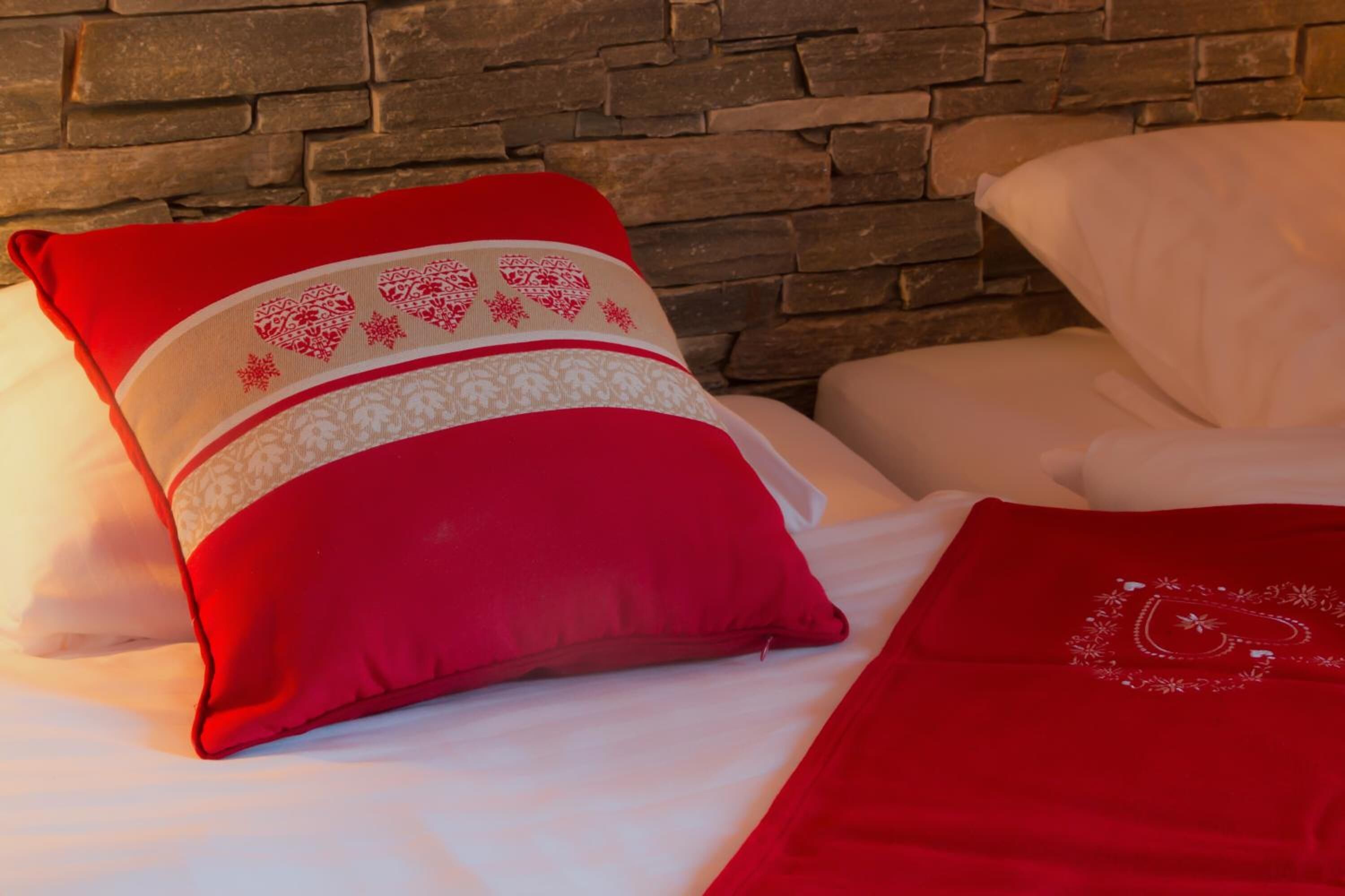A bed with a red pillow and a red blanket, a white pillow, and a stone wall in the background.