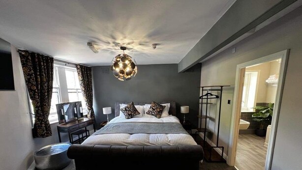 A modern bedroom with a large bed, bedside tables, a hanging light fixture, and a view of the bathroom through an open door. - Elephant & Castle (Holmfirth)