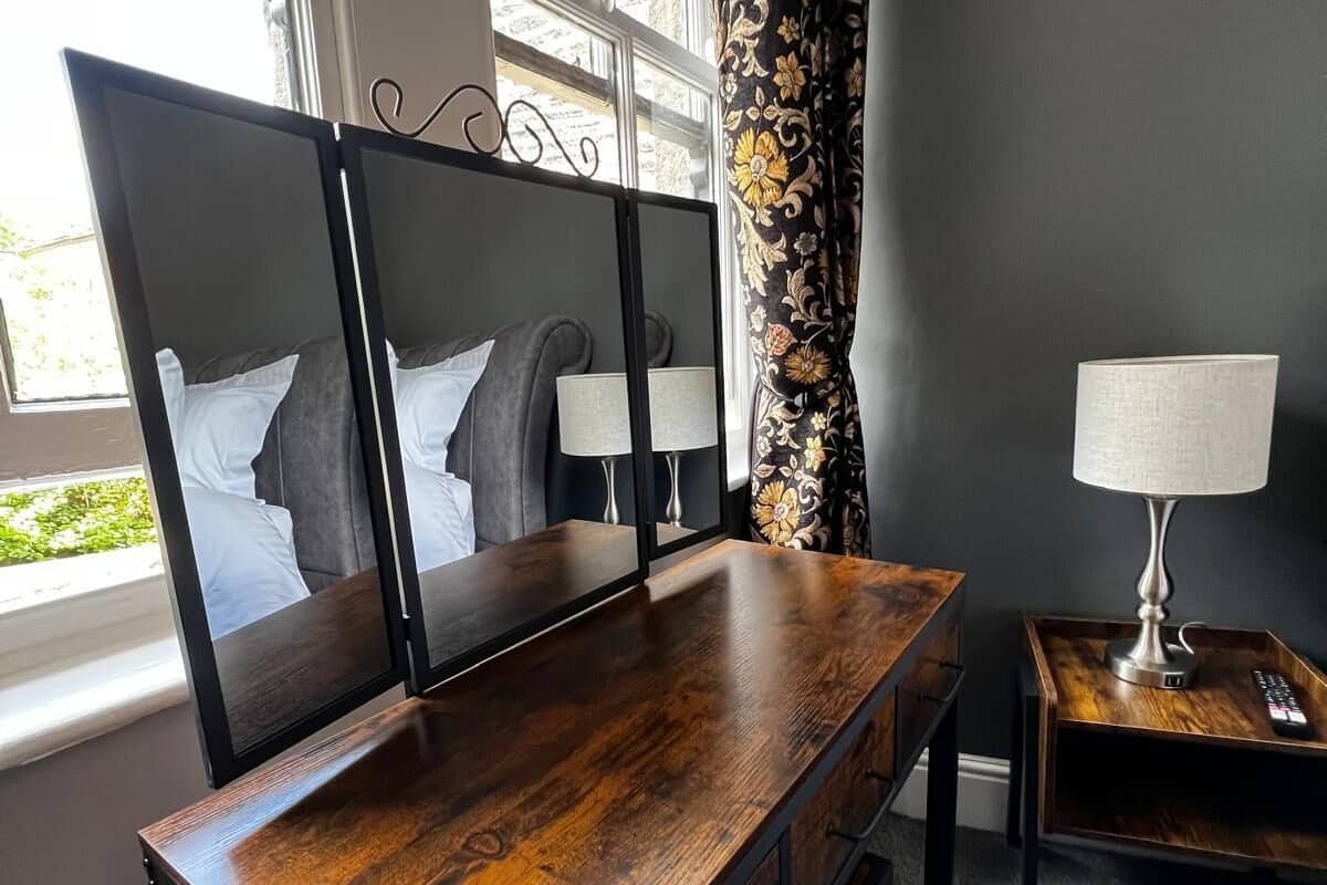 A room with a wooden desk, a TV stand with two screens, a lamp, and a window with curtains.