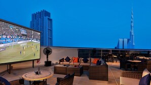 A rooftop lounge with wicker furniture, a large TV screen, and a view of the Burj Khalifa. - Carlton Downtown (Dubai)