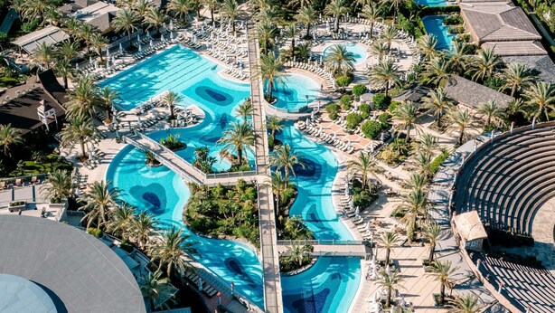 An aerial view of a resort with a large central swimming pool, palm trees, and a beach area. - Royal Wings Hotel - All Inclusive (Antalya)