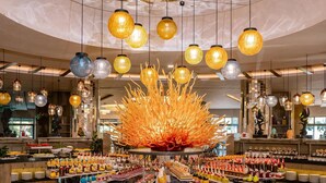A luxurious lobby with a central round wooden table surrounded by food and drink stations, overhead hanging lights, and a large reflective ceiling. - Royal Wings Hotel - All Inclusive (Antalya)