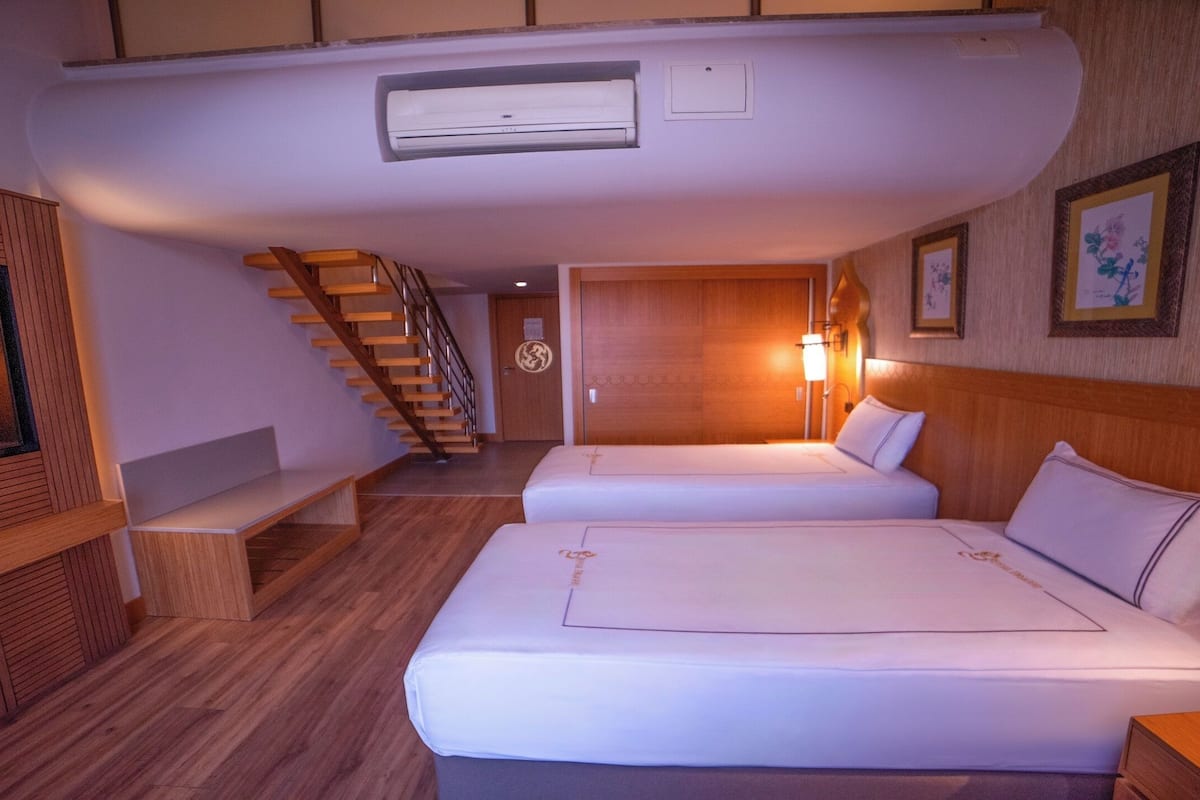 A hotel room with two beds, a wooden bench, a staircase, and a wall-mounted air conditioner.