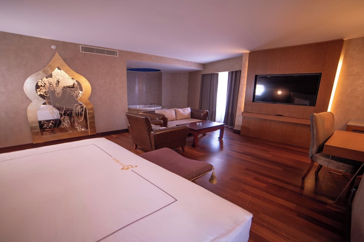 A hotel room with a large bed, a seating area with a sofa and chairs, a TV mounted on the wall, and a bathtub in the background.