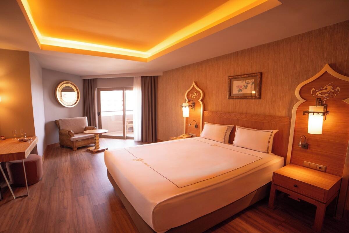 A hotel room with a large bed, a desk with a chair, a round mirror, and a window with curtains.