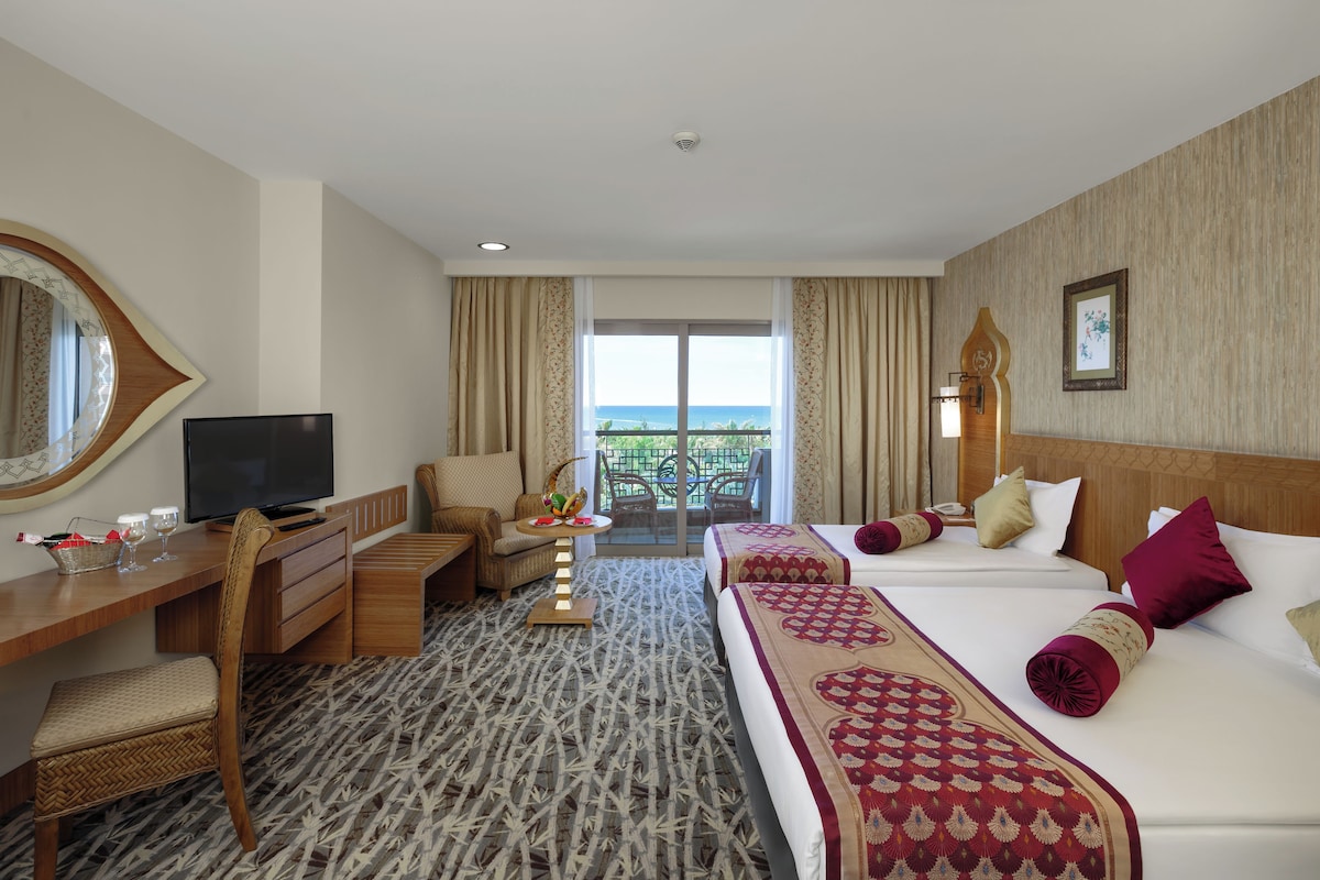 Ein Hotelzimmer mit Bett, Schreibtisch, Fernseher, Sessel, einem kleinen Tisch mit Lampe und Blick ins Freie.