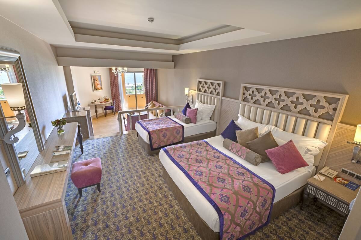 A hotel room with a large bed, a smaller bed, a desk, and a chair. There is a window with curtains, a mirror, and a decorative headboard.
