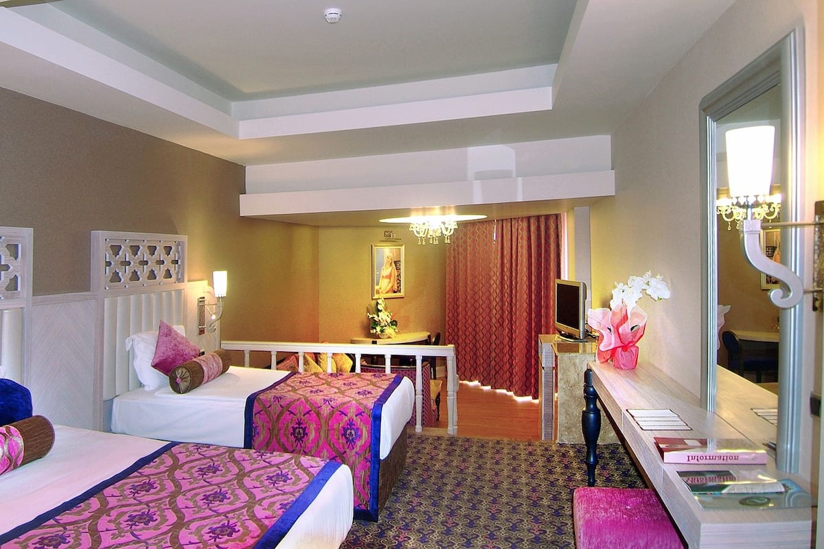 A hotel room with two beds, a desk, a mirror, and a chandelier.
