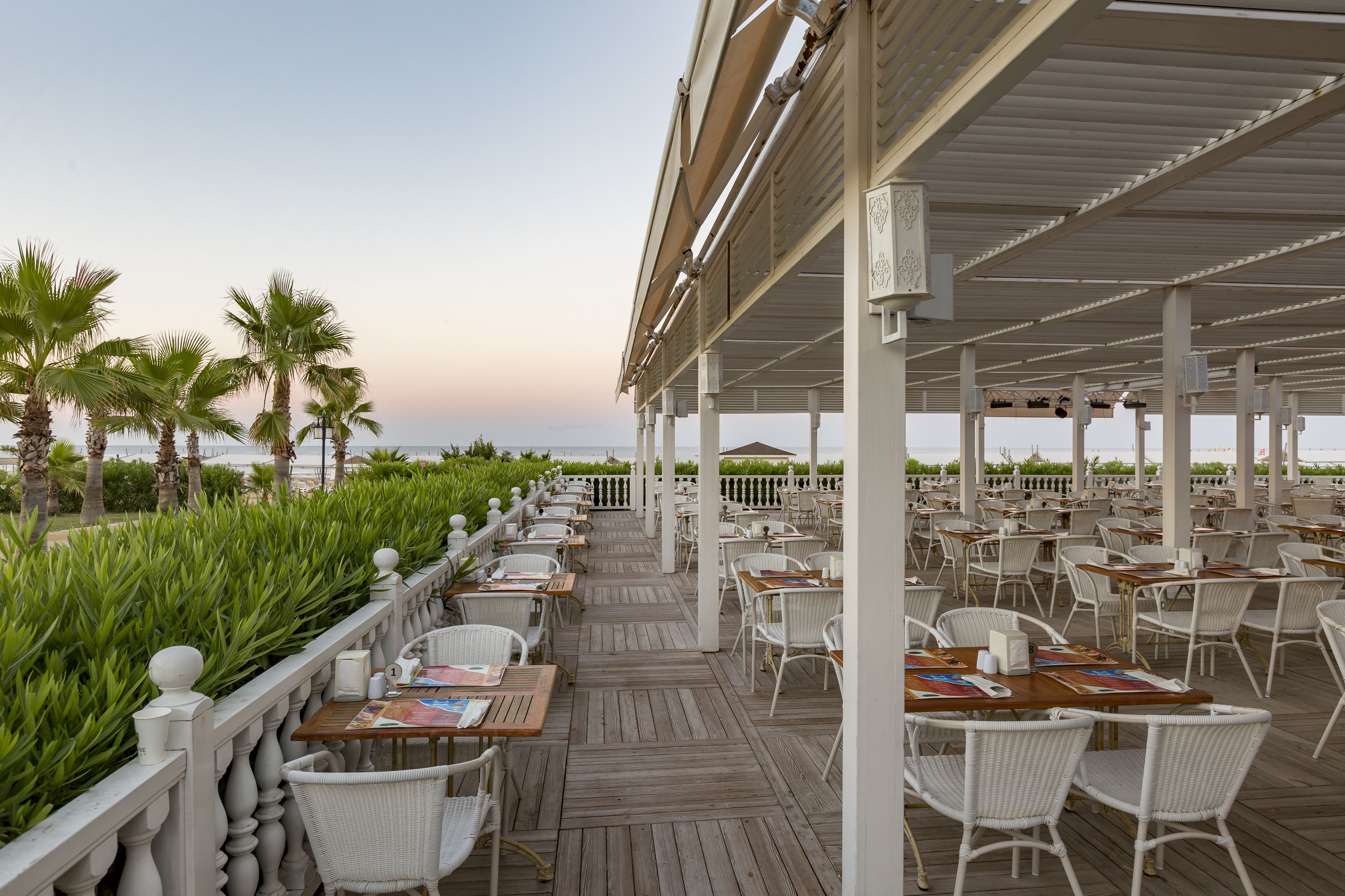 A covered outdoor dining area with white wicker chairs and tables, overlooking a beach with palm trees.