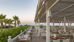 A covered outdoor dining area with white wicker chairs and tables, overlooking a beach with palm trees. - Royal Alhambra Palace – All Inclusive (Manavgat)