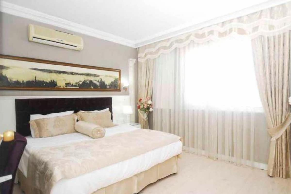 A bedroom with a bed, a painting, a window with curtains, and a vase with flowers.