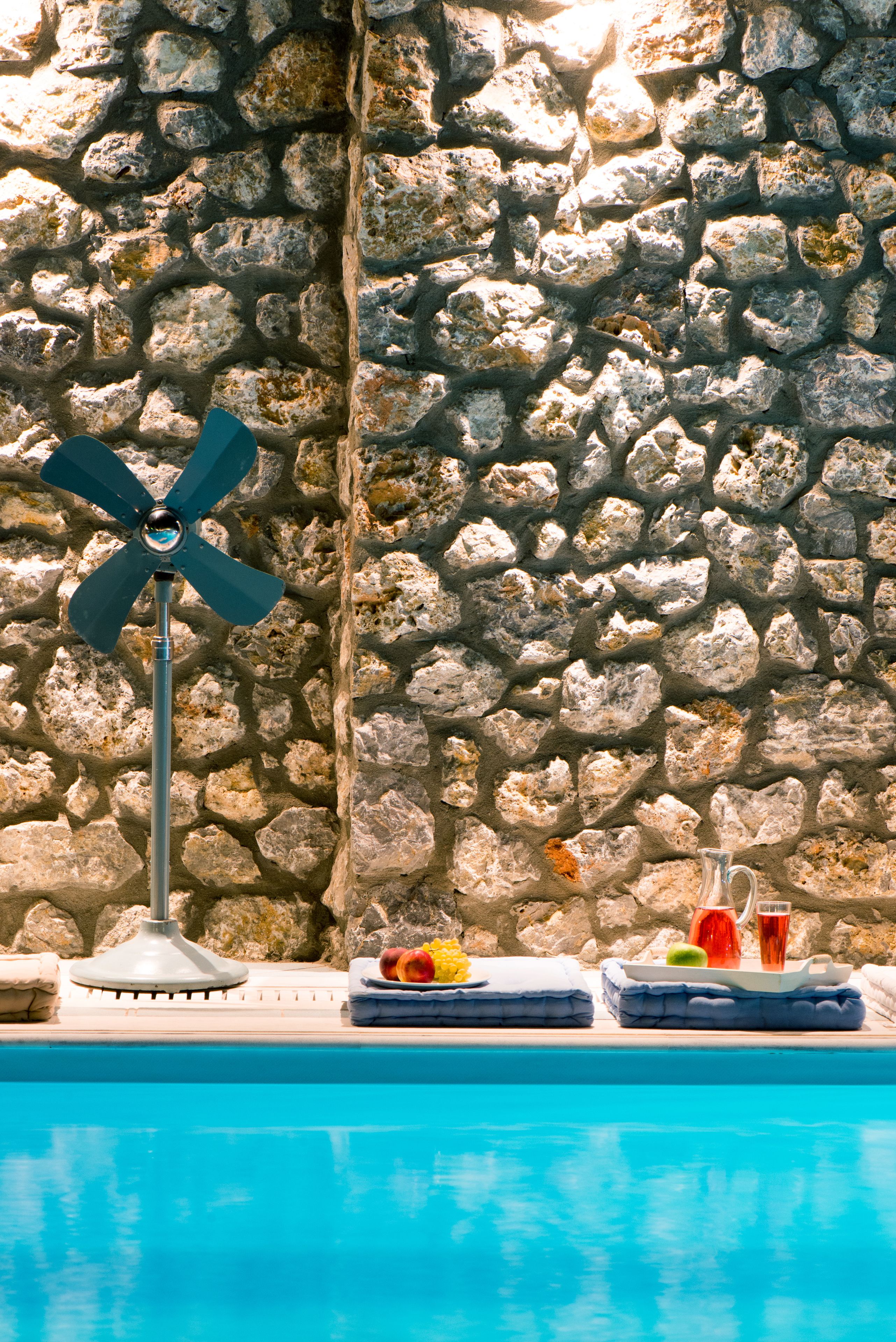 A pool area with a stone wall, a ceiling fan, and a tray with fruits and drinks.