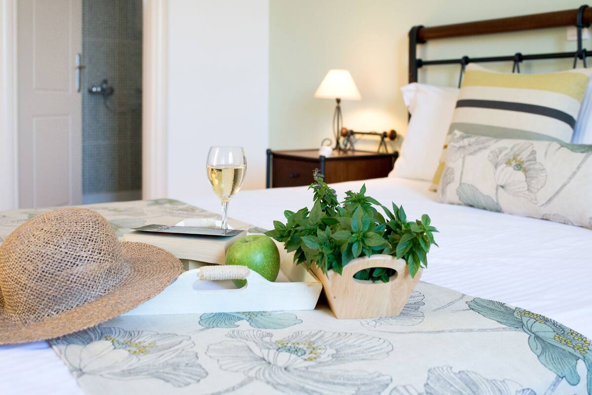 A neatly made bed with a floral bedspread, a green apple, a glass of white wine, a straw hat, and a wooden planter with green leaves.