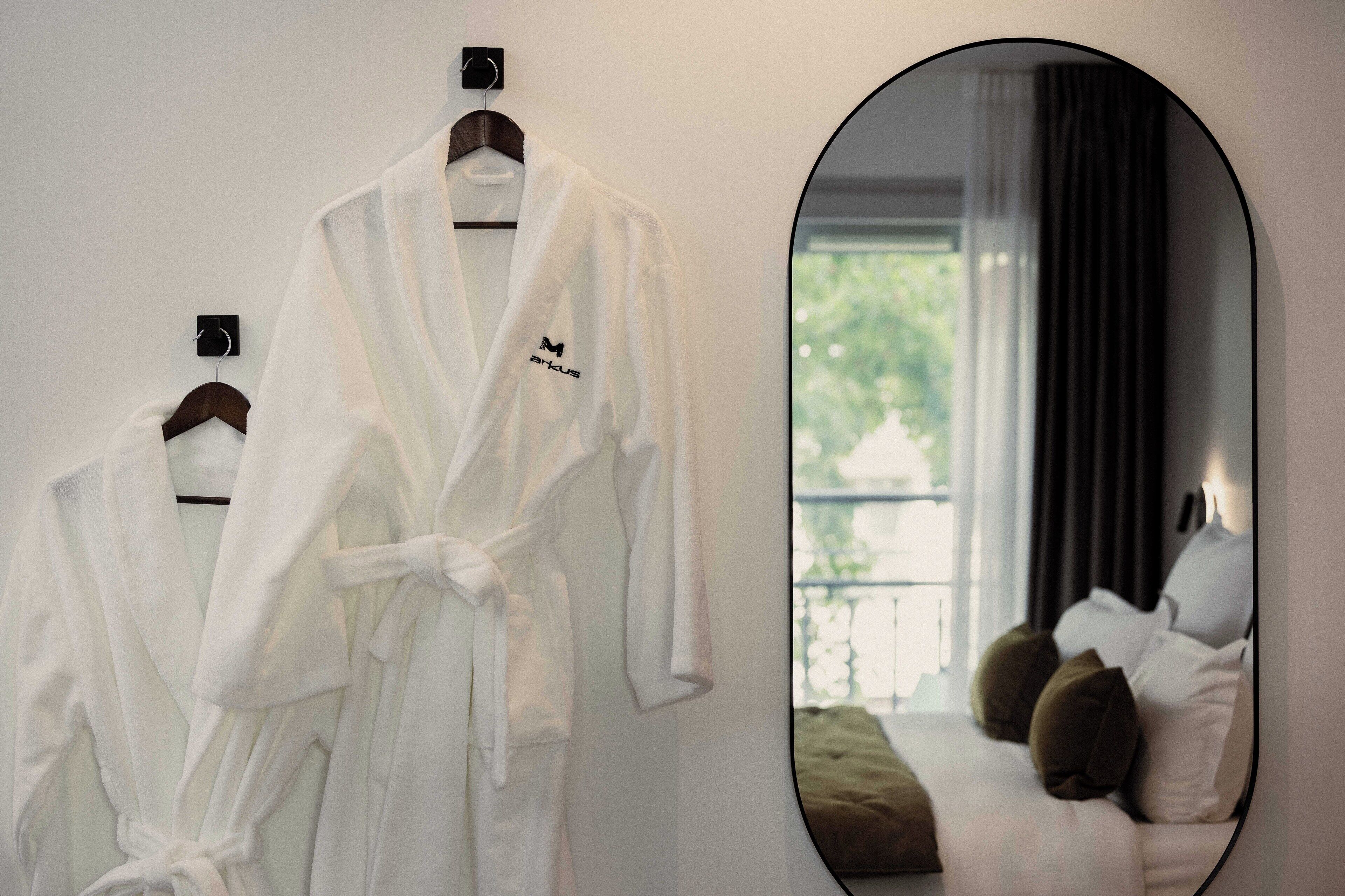 Una bata blanca colgada en una percha de madera, un espejo redondo que refleja una cama con almohadas, y una ventana con cortinas.