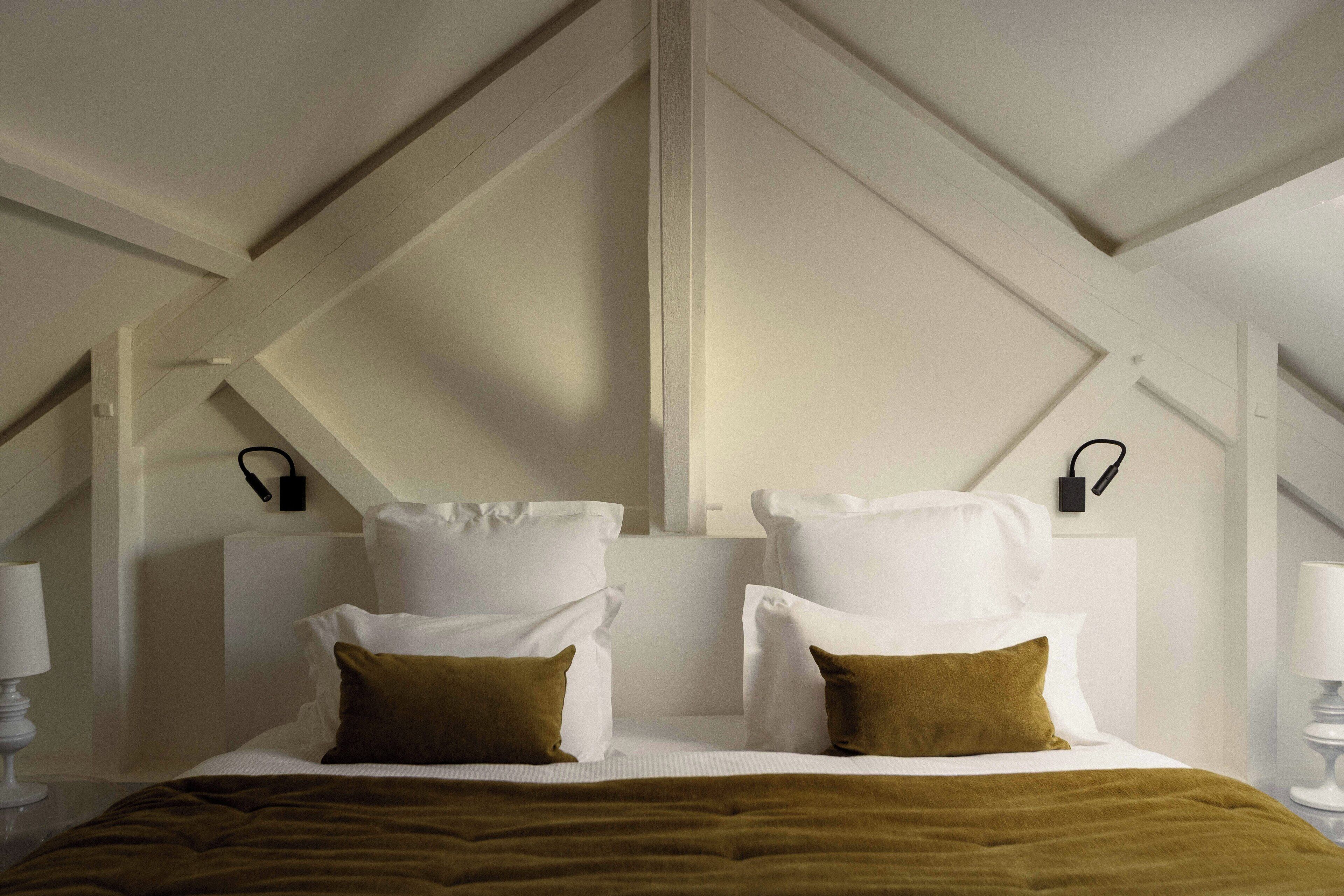 Un dormitorio con techo alto, paredes blancas y techo inclinado. Hay dos mesitas de noche con lámparas, una cama con ropa de cama blanca y almohadas marrones, y una manta marrón.