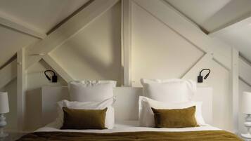 Un dormitorio con techo alto, paredes blancas y techo inclinado. Hay dos mesitas de noche con lámparas, una cama con ropa de cama blanca y almohadas marrones, y una manta marrón.