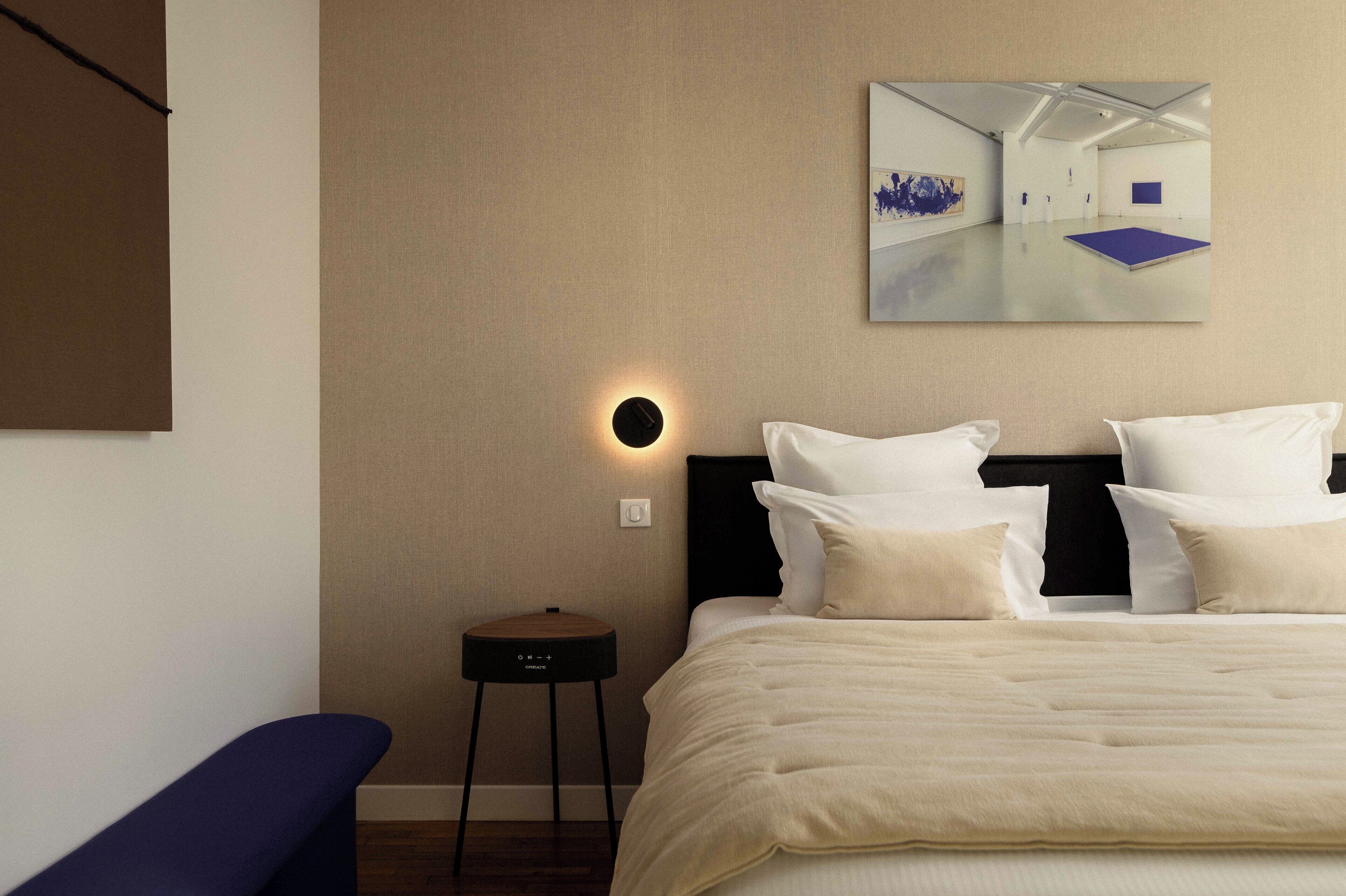 Moderni hotellihuone, jossa on suuri sänky, pieni sivupöytä, penkki ja seinälle kiinnitetty taideteos.