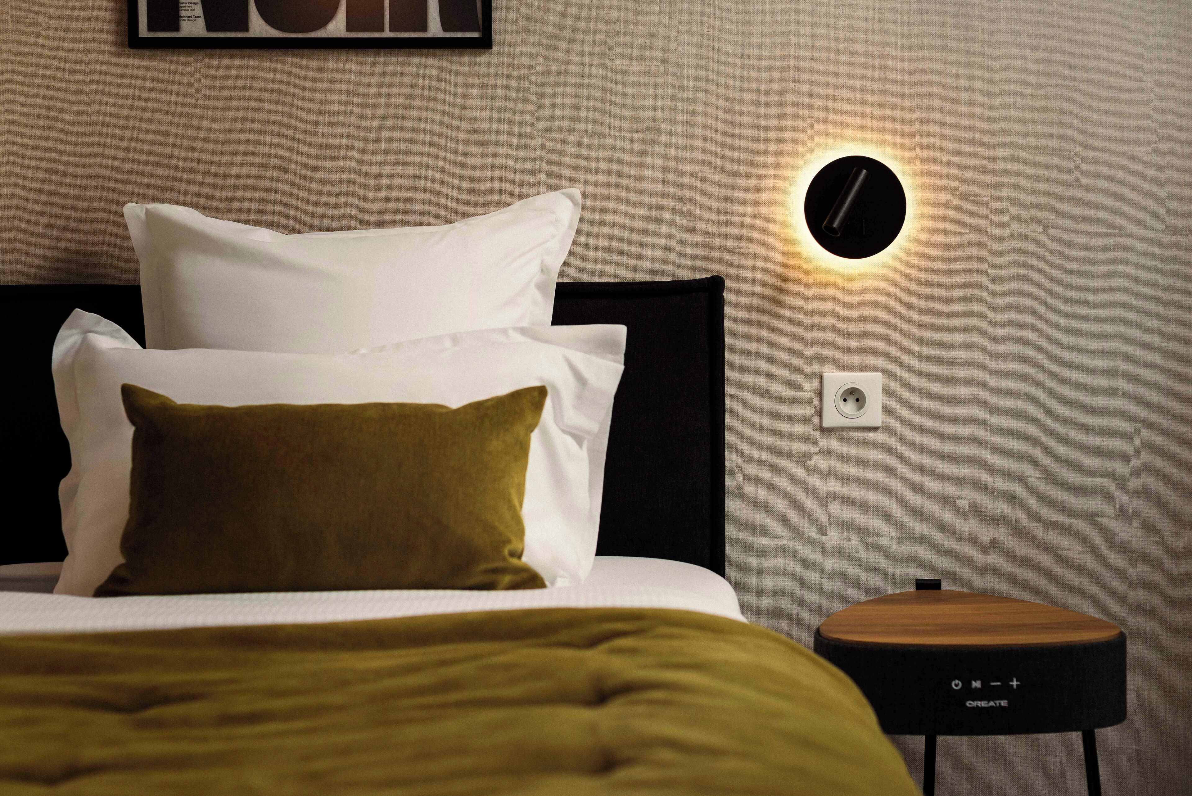 A neatly made bed with white and olive green pillows, a round wall-mounted light, and a small wooden side table with a black base.