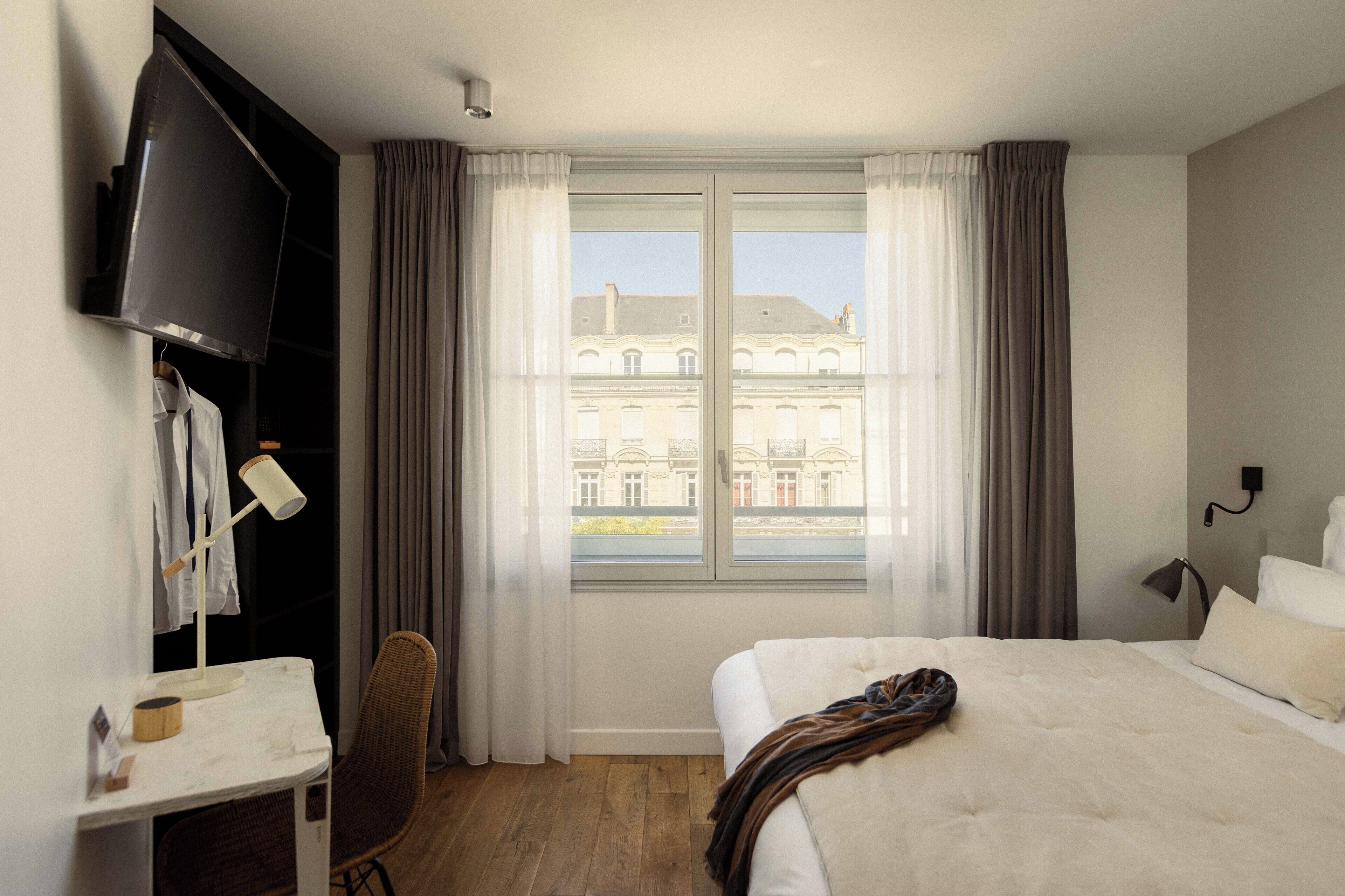 Una habitación de hotel con cama, escritorio, silla, televisor y ventana con cortinas.