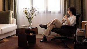 A woman sitting in a leather chair in a well-lit room with a white sofa, a small table with a vase of flowers, and a window with curtains. - Markus (Angers)