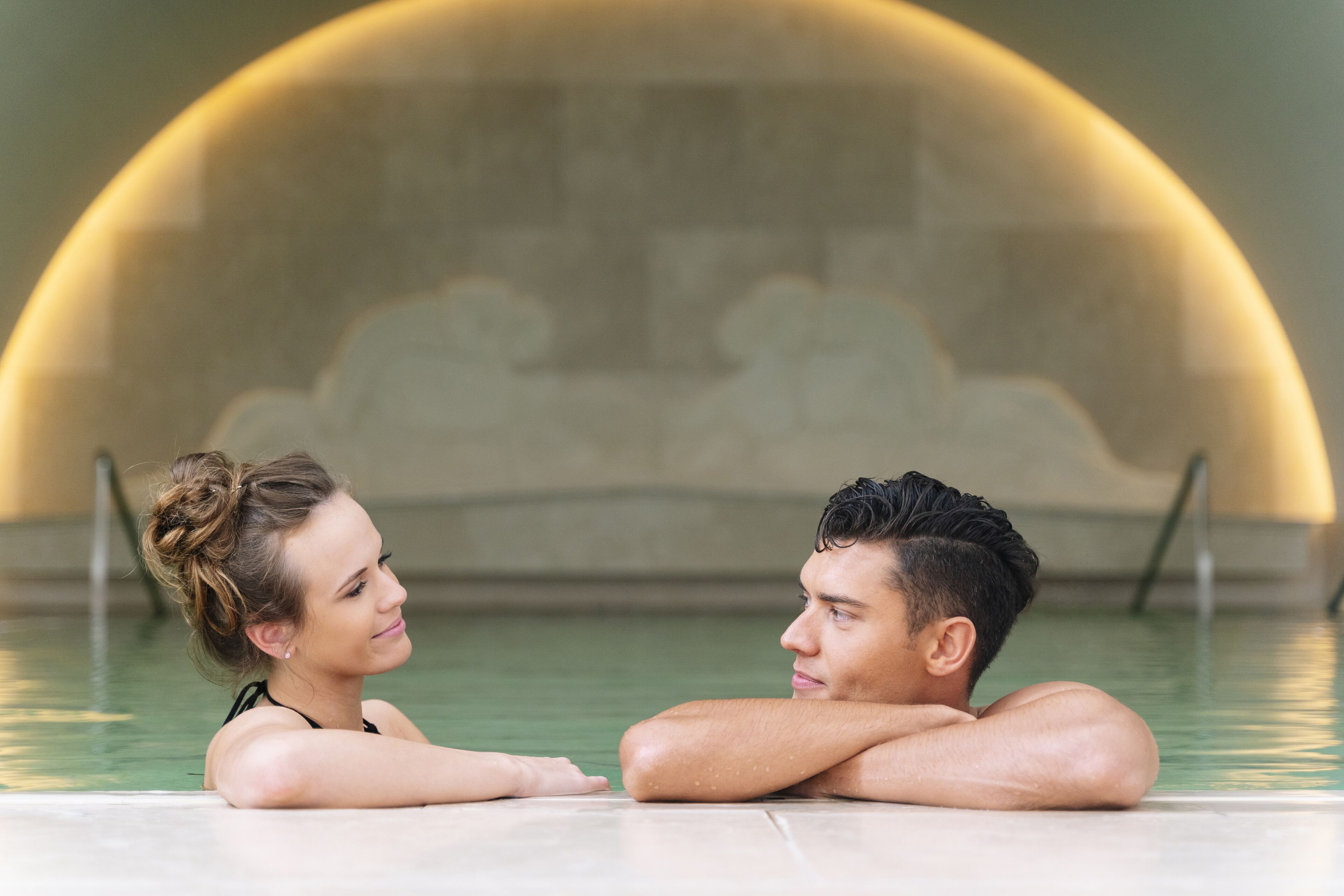 A couple in a swimming pool with a stone wall and a curved arch in the background.