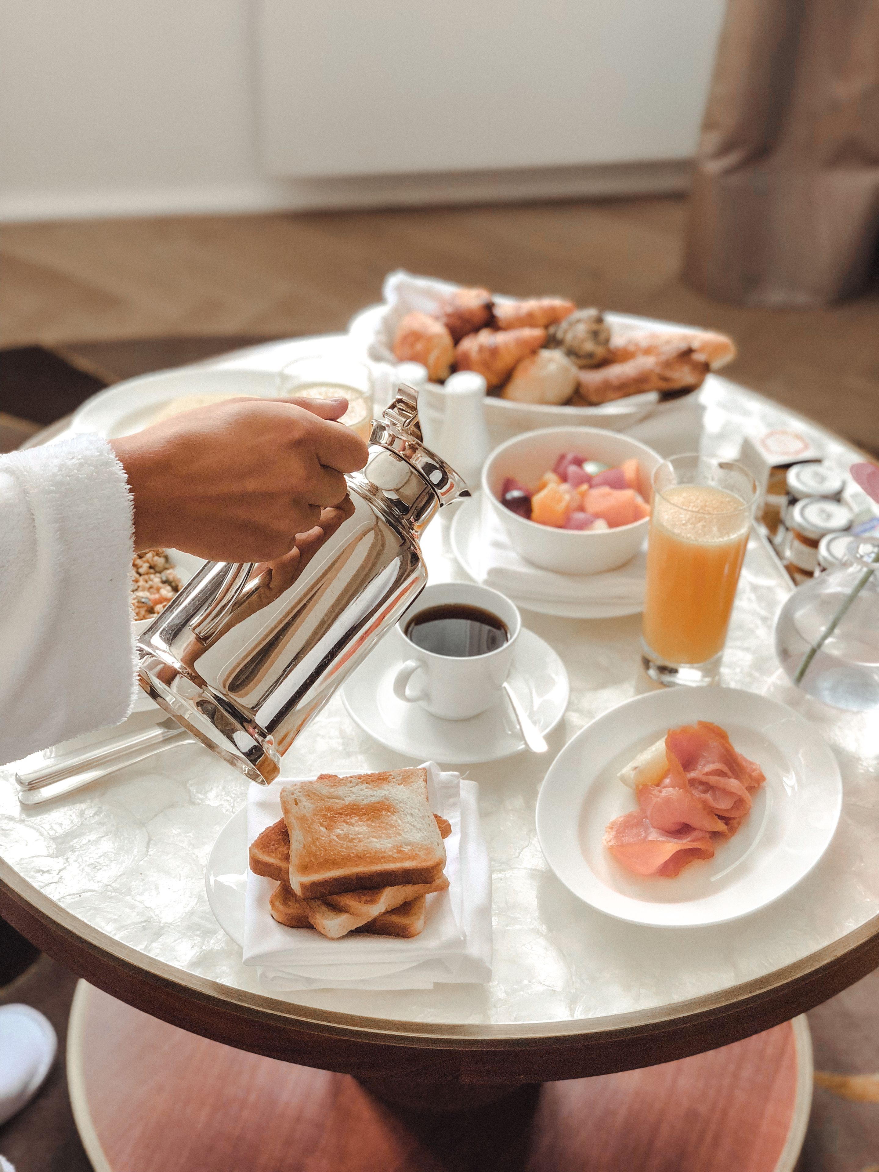 A breakfast setting with a coffee pot, croissants, fruit, and ham on a table.