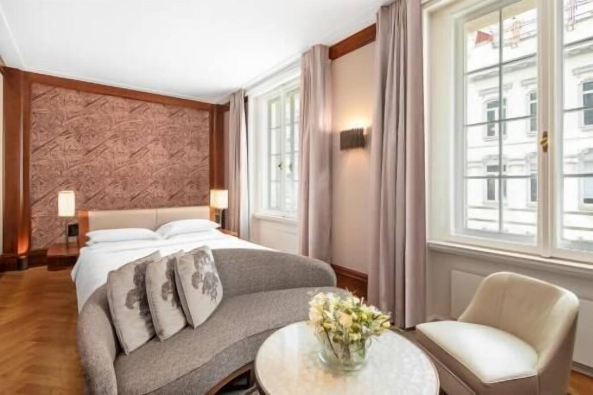 A hotel room with a bed, a sofa, a chair, a table with flowers, and large windows.