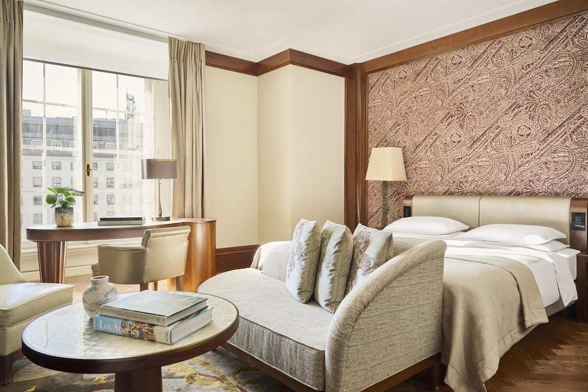 Een hotelkamer met een groot raam, een bank, een nachtkastje, een lamp en een bekeken hoofdeinde.