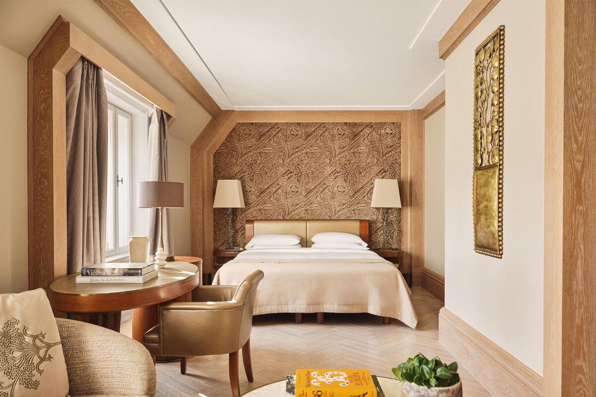 Een hotelkamer met een bed, twee bedlampjes, een ronde tafel met een lamp, een stoel, een klein tafeltje met boeken en een decoratief paneel aan de muur.