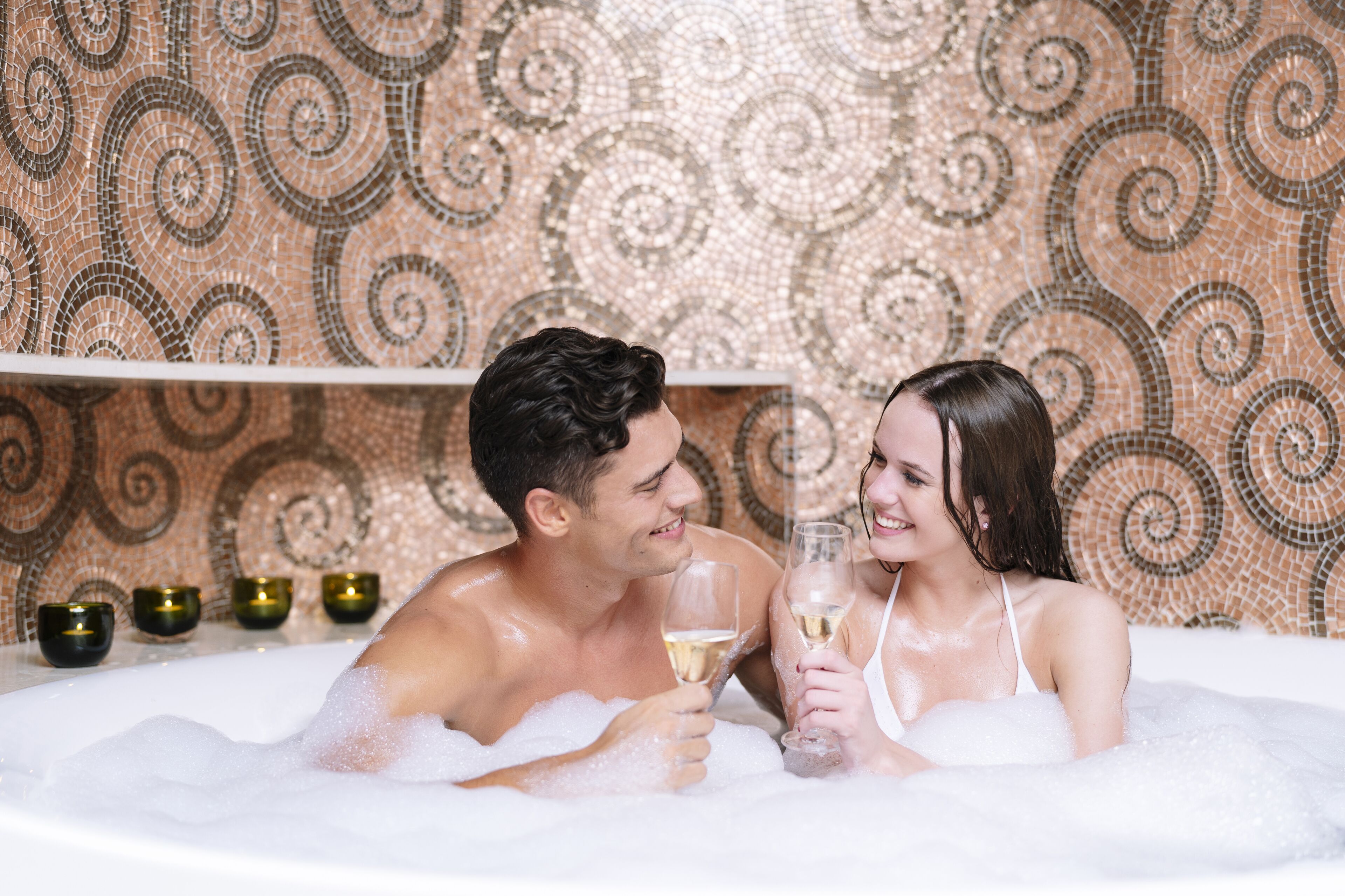 Un couple se détend dans un bain à remous, avec un mur décoré de mosaïques en arrière-plan.