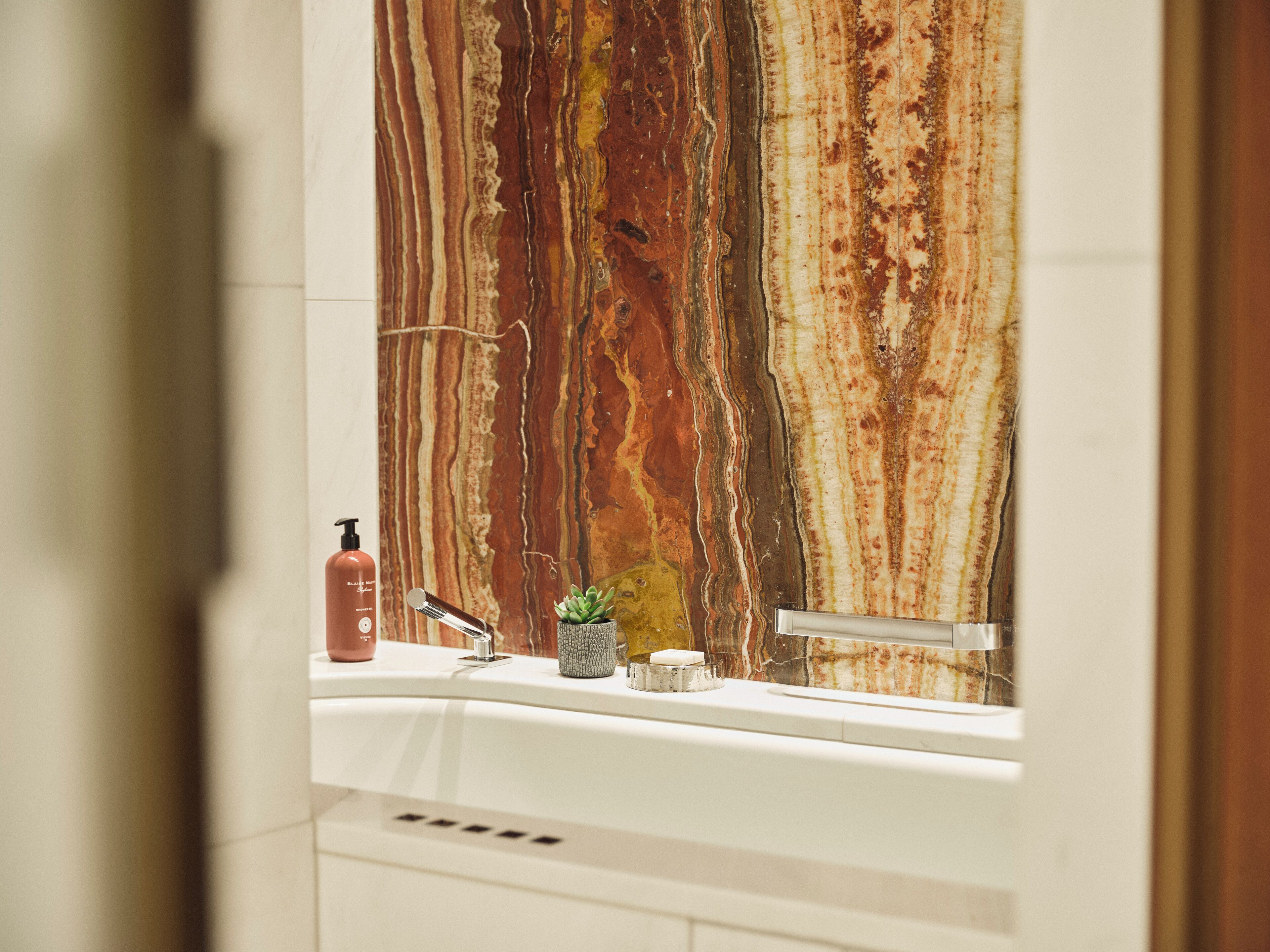 Une salle de bain avec un pare-baignoire en marbre, un lavabo, un robinet, un distributeur de savon et une petite plante.