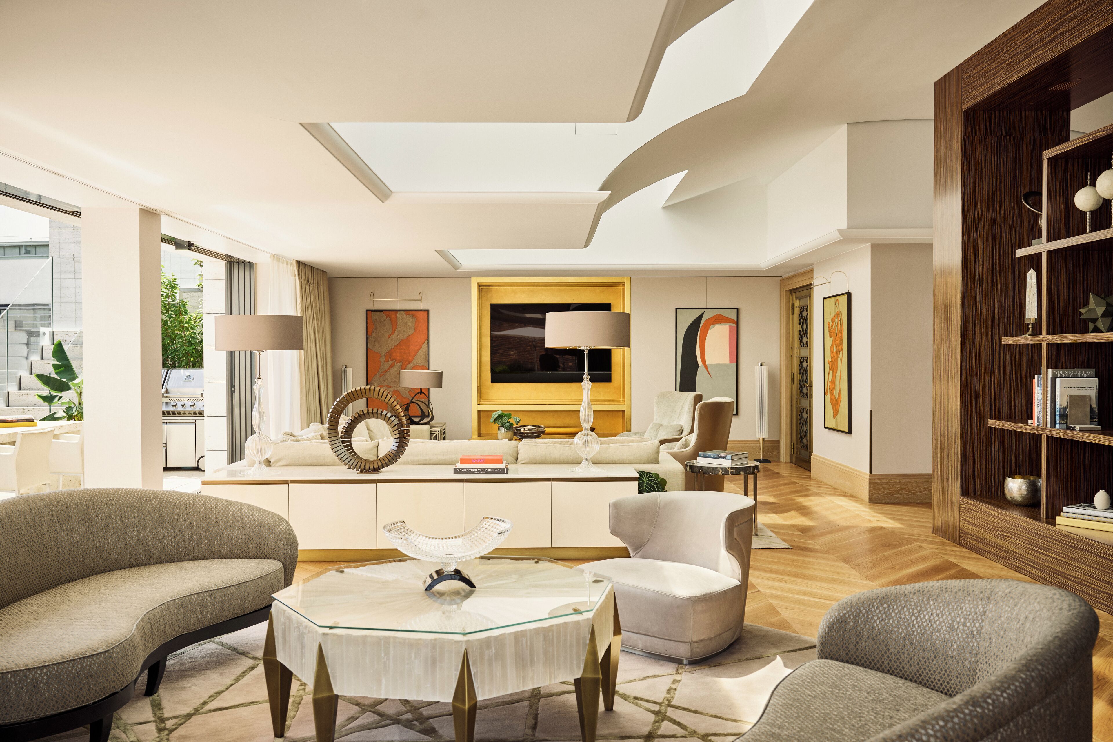 A spacious living room with a sofa, armchairs, a glass coffee table, and a wooden bookshelf.