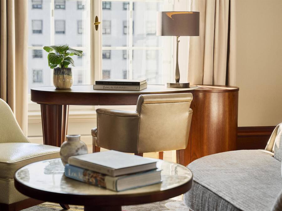 A room with a leather armchair, a round table with books, a desk with a lamp, and a potted plant.
