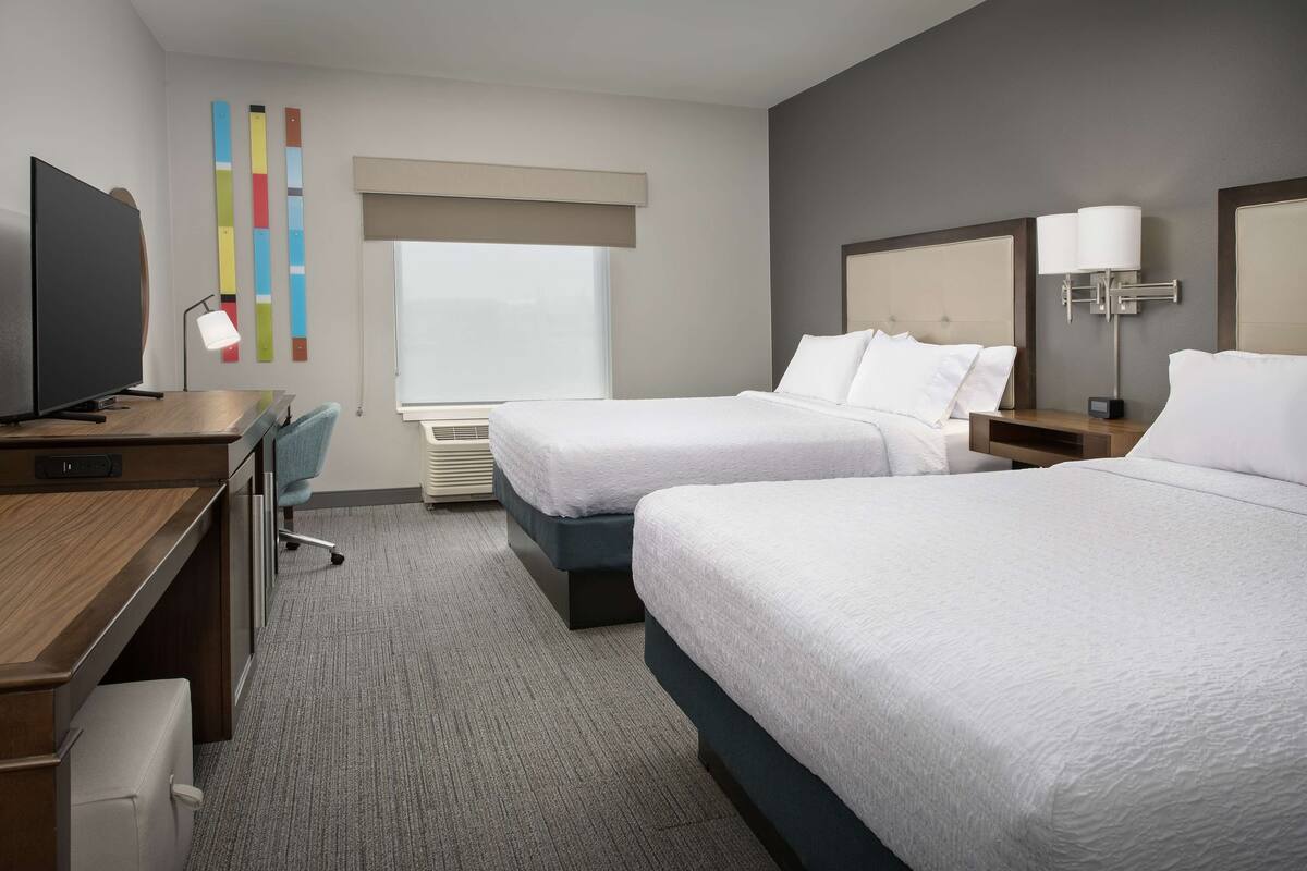 A hotel room with two beds, a desk with a computer, a TV, and a window with blinds.