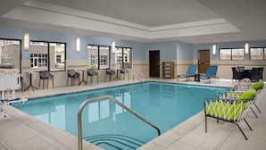 A hotel indoor pool area with a clear pool, lounge chairs, and a seating area with tables and chairs.