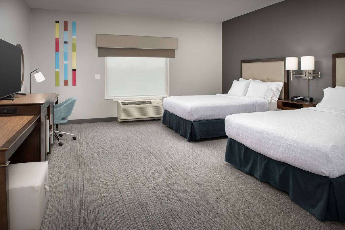 A hotel room with two beds, a desk with a computer, a chair, a TV, and a window with blinds.