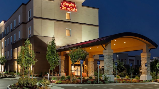 A Hampton Inn & Suites hotel with a modern design, featuring a prominent neon sign, a covered entrance, and landscaped areas with potted plants.