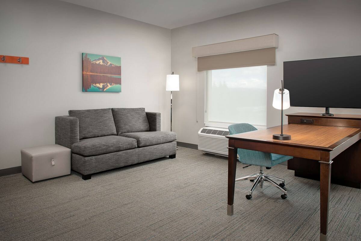 A hotel room with a gray sofa, a wooden desk, a chair, a small ottoman, a large window with a view, a painting on the wall, and a flat-screen TV.