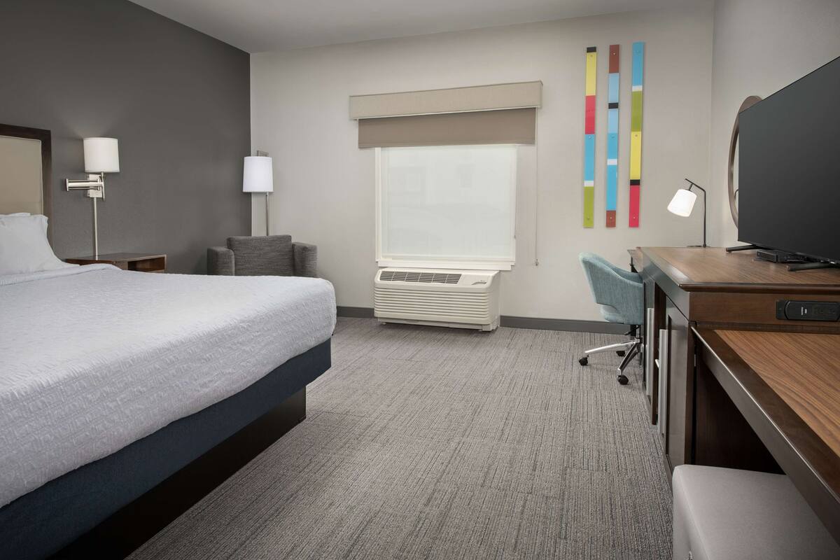 A hotel room with a large bed, a desk with a computer, a TV, a window with blinds, and a wall-mounted air conditioner.