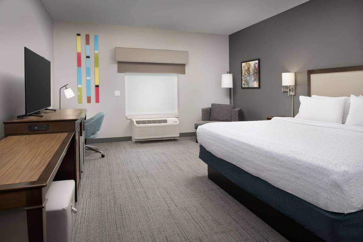 A hotel room with a bed, desk, chair, air conditioning unit, and a window with blinds.