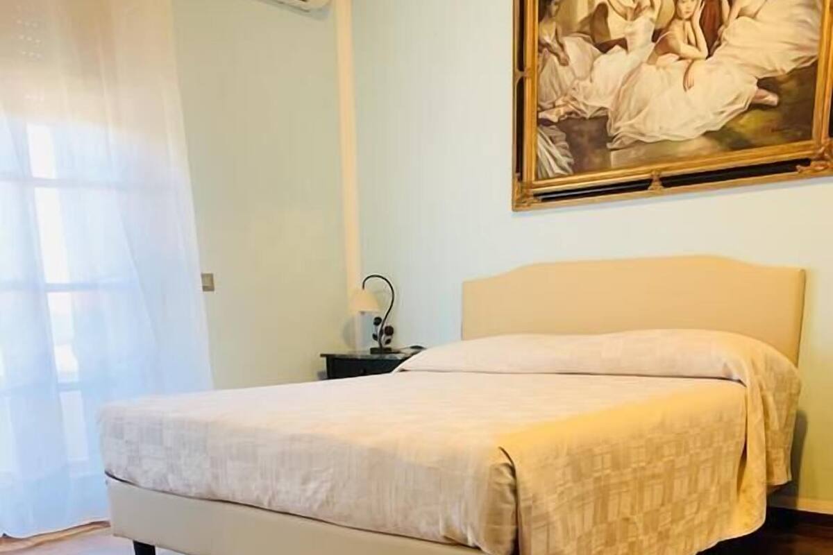 A bedroom with a bed, wooden parquet flooring, a nightstand, a lamp, and a painting on the wall.