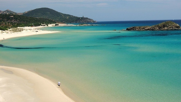 Uma praia com águas turquesa cristalinas, areal arenoso e uma pessoa caminhando pela orla.