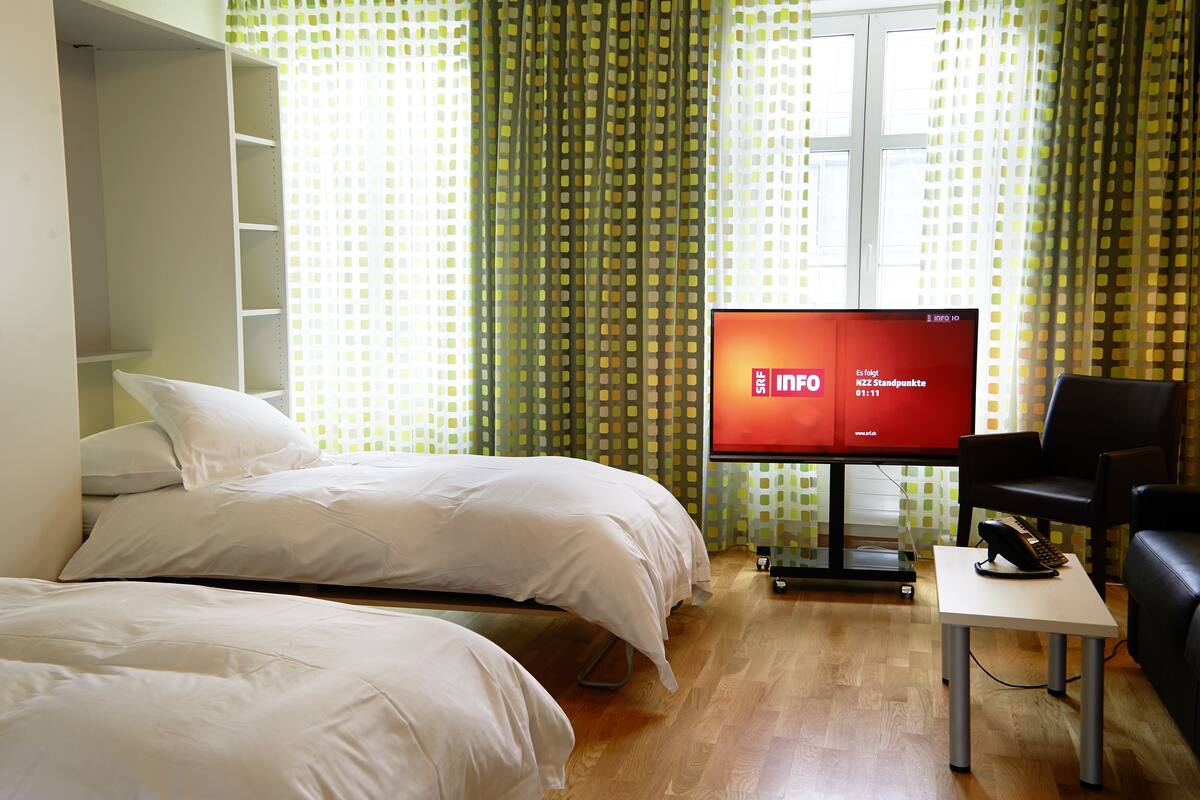 Ein Hotelzimmer mit zwei Betten, einem Schreibtisch mit Stuhl, einem Fernseher und einem Fenster mit Vorhängen.