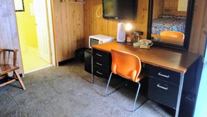 Una habitación compacta con un escritorio de madera, una silla, un televisor montado en la pared, un espejo y una cama con una colcha colorida.