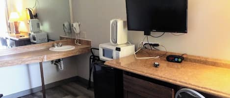 A hotel room with a wooden counter, a sink, a microwave, a hairdryer, a television, and a chair.