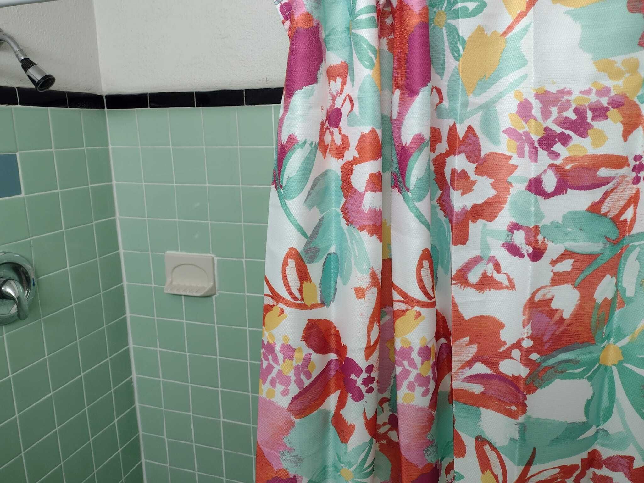 A bathroom with green tiles, a shower curtain with a floral pattern, and a visible showerhead.