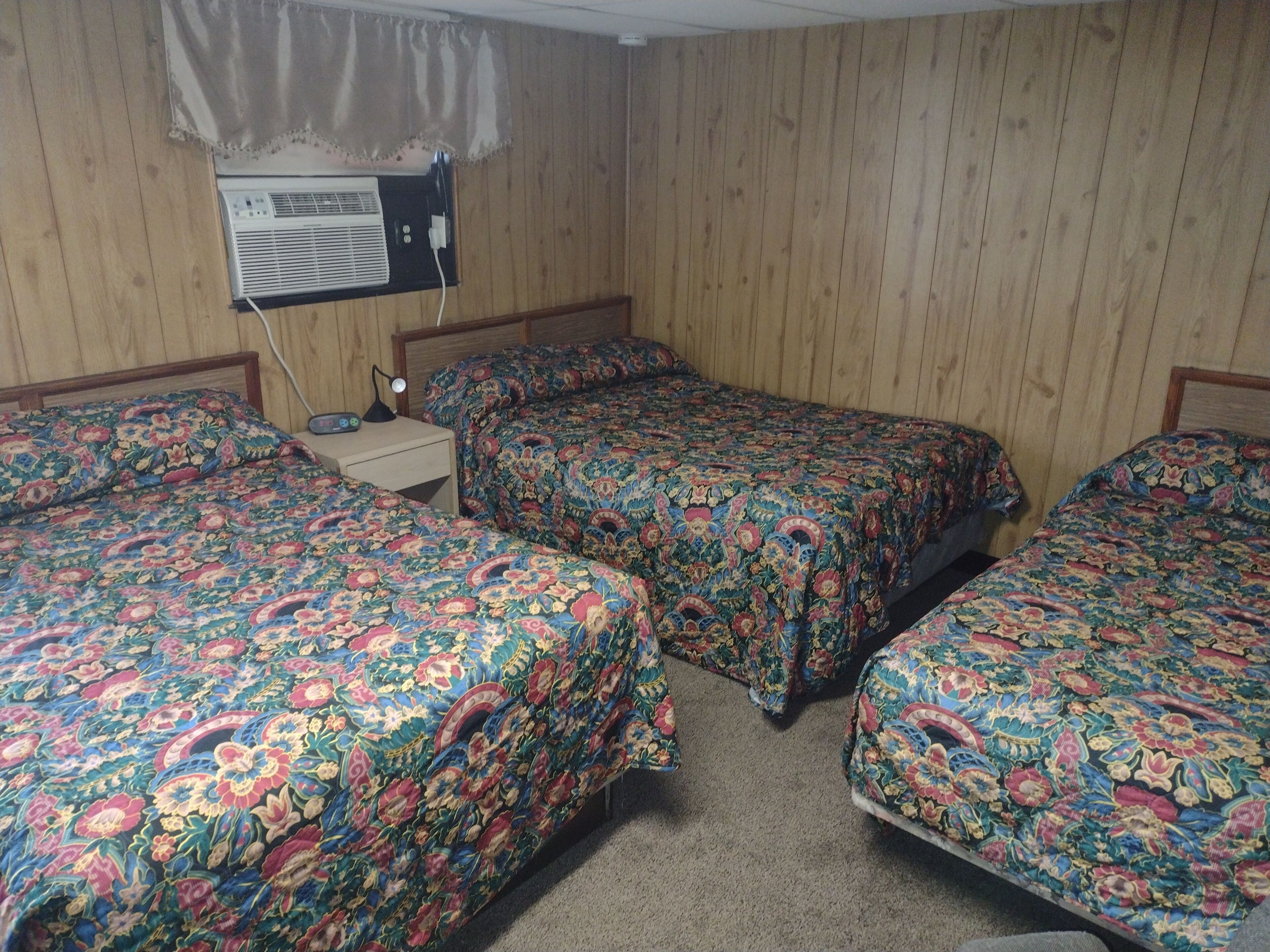 A room with three beds, each with a floral-patterned bedspread. There is a wall-mounted air conditioner and a nightstand with a lamp.